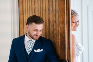 Groom in blue suit smiles while bride peeks around wooden door during wedding first look.