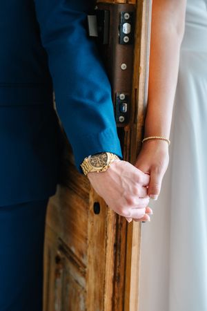 Close-up of bride and groom holding hands, groom wearing gold watch, near wooden door.