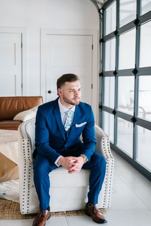 Groom in navy blue three-piece suit sitting in white tufted chair by large window on wedding day.