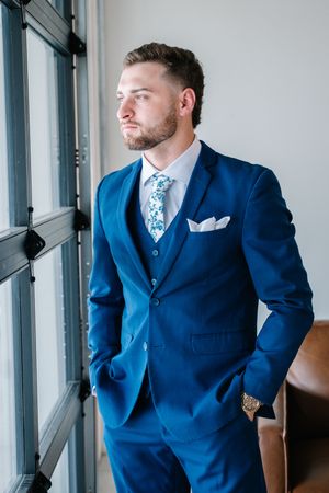 Handsome groom wearing navy blue three-piece suit with floral tie standing by window with hands in pockets.