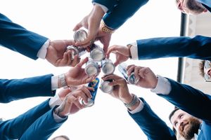 Groomsmen toasting with silver flasks in a circle, view from below showing blue suits and watches.