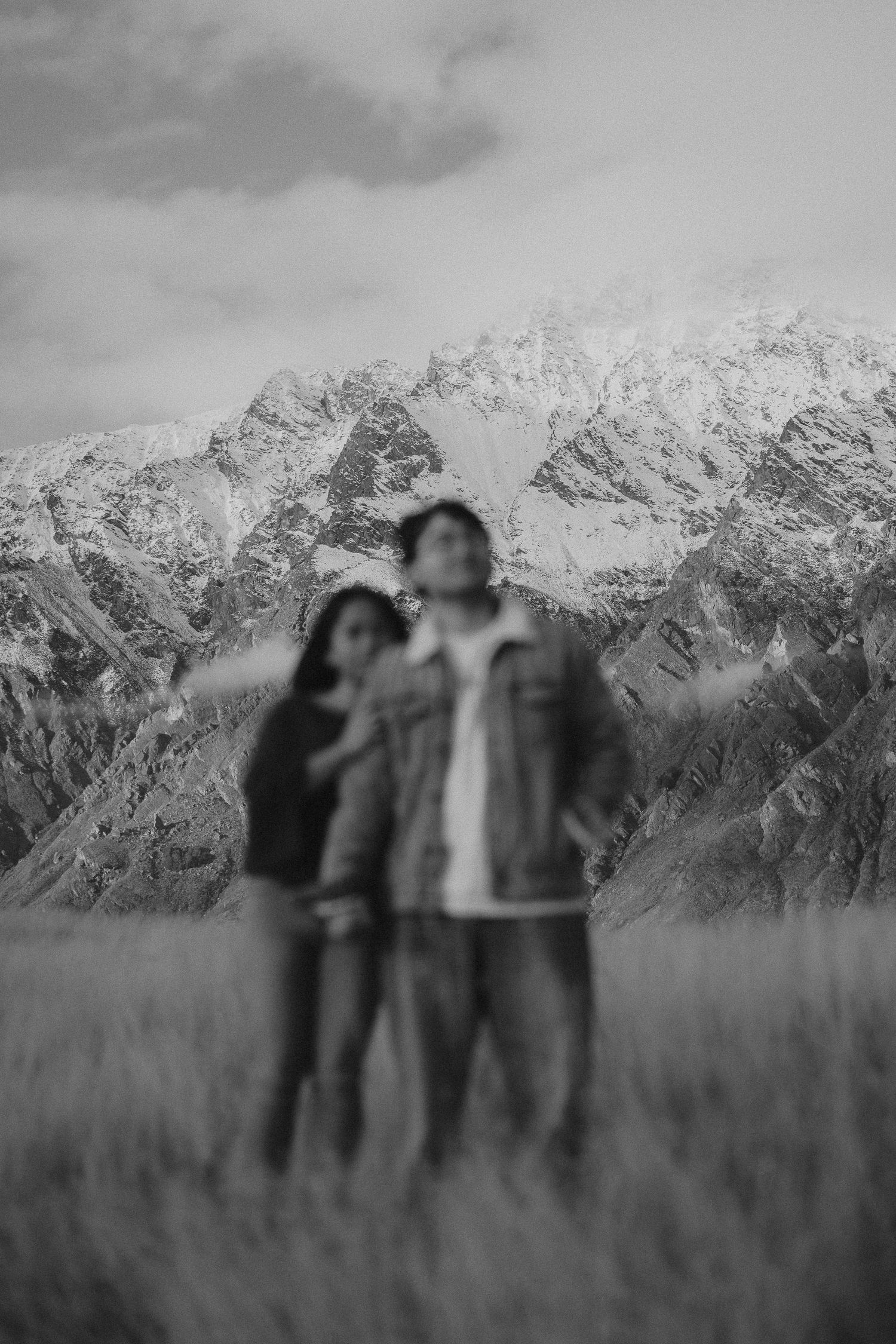 Couple dancing joyfully on a grassy hillside with snow-capped mountains and lake in background.