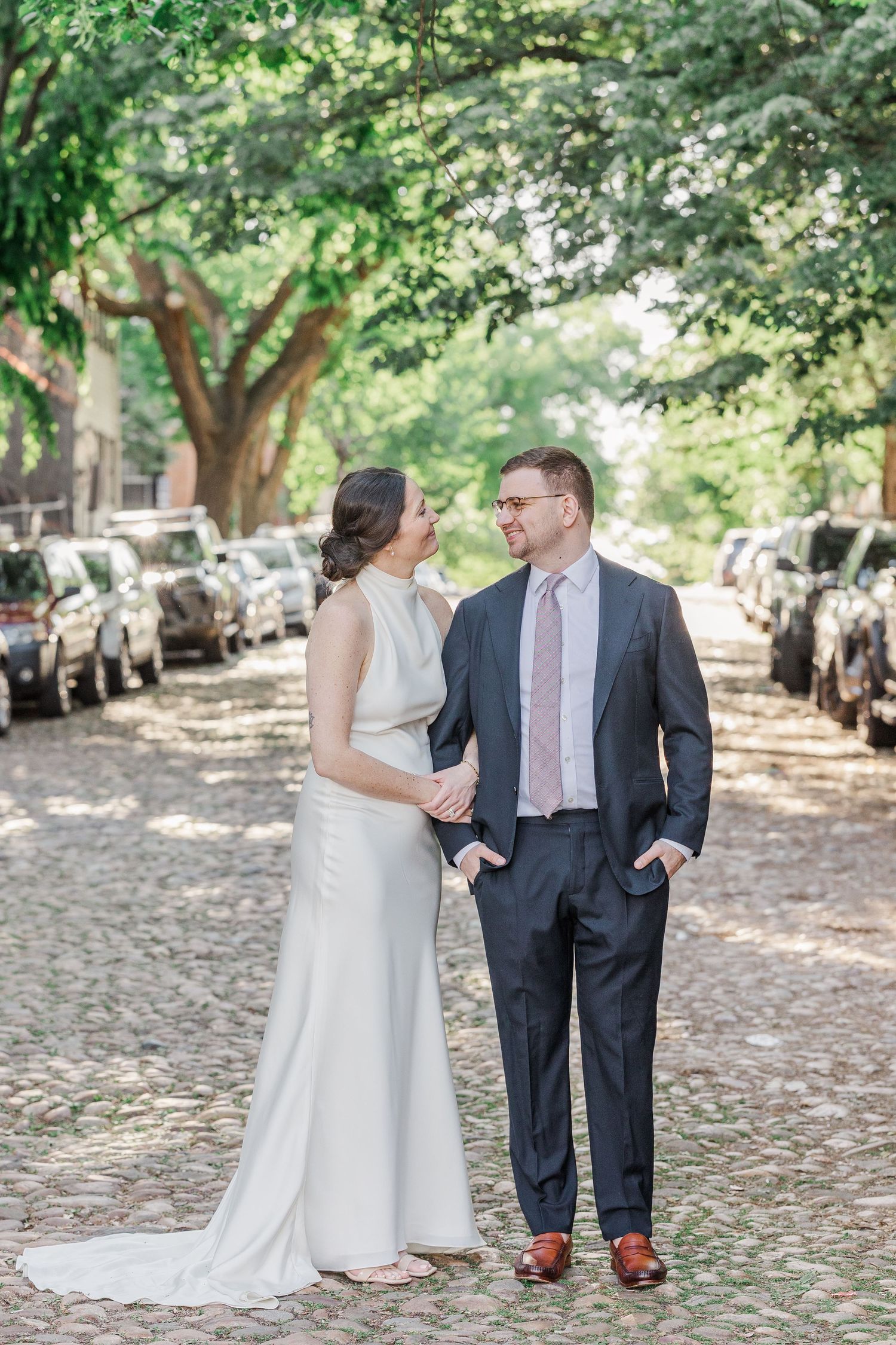 A bride and groom walk hand-in-hand on a cobblestone street lined with trees and parked cars, smiling at each other.
