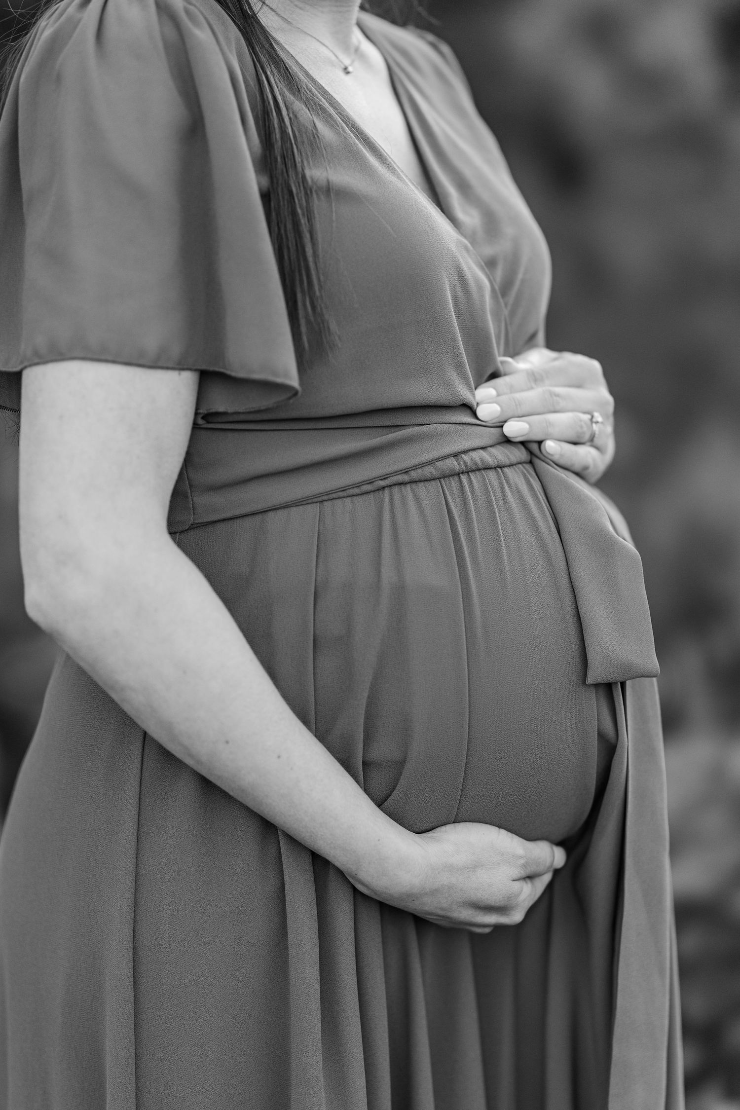 Black and white photo of a pregnant woman in a flowy dress cradling her baby bump with both hands outdoors.