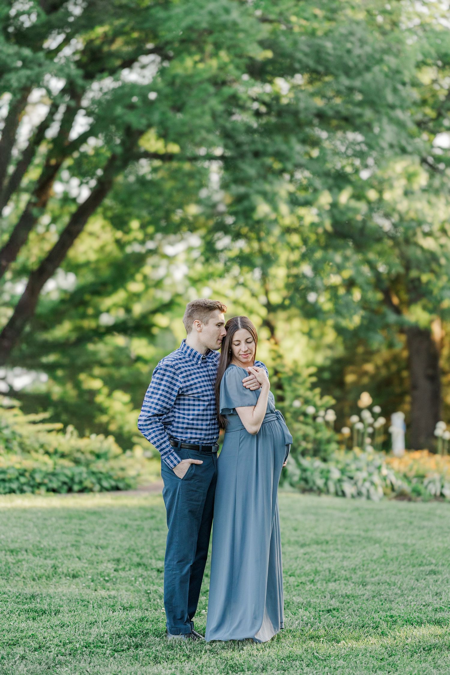 A couple embraces in a lush green park, woman in flowing blue dress, man in plaid shirt, sharing a tender moment.