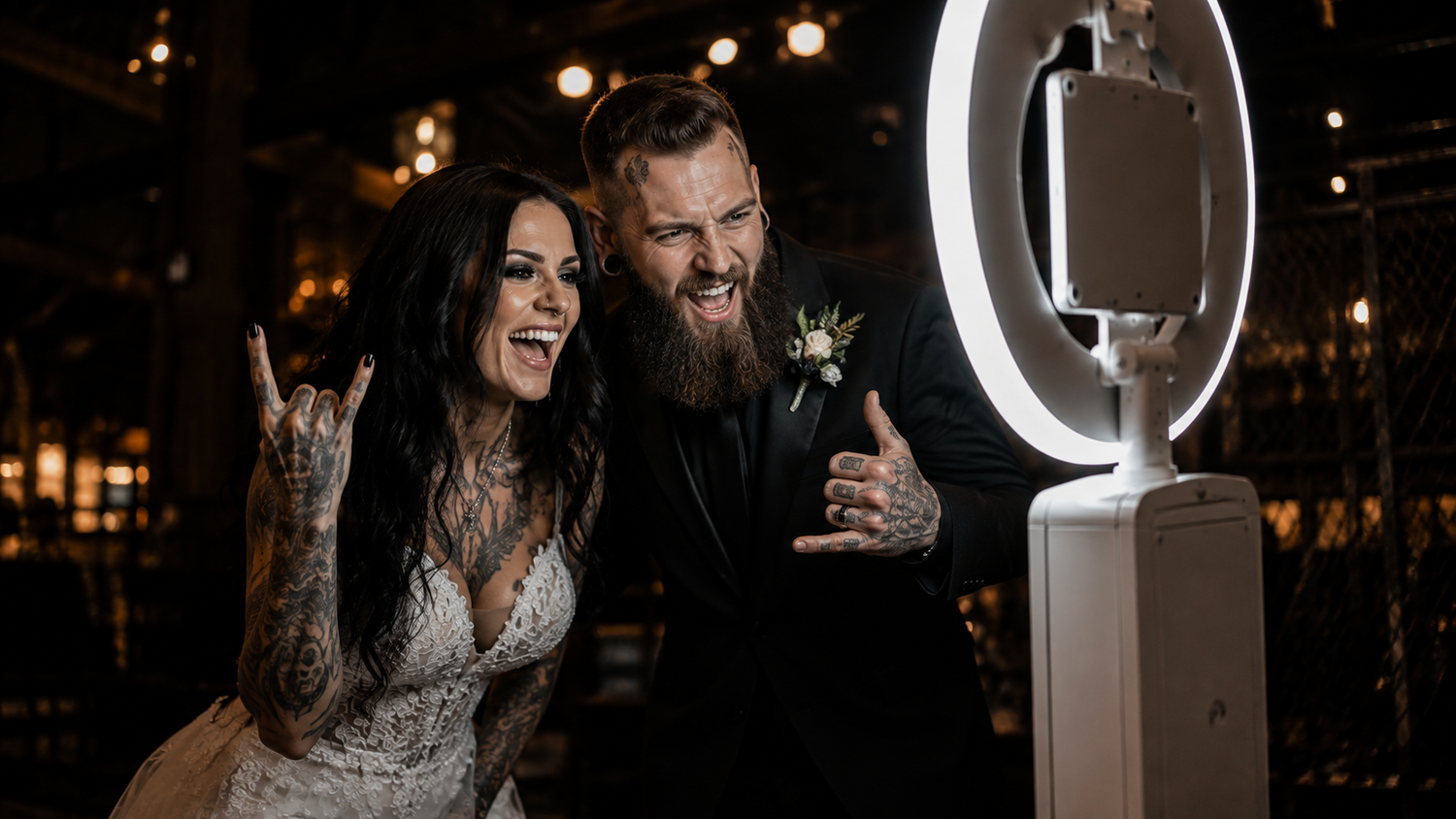 Tattooed bride and groom pose playfully beside a ring light photo booth at their wedding reception.