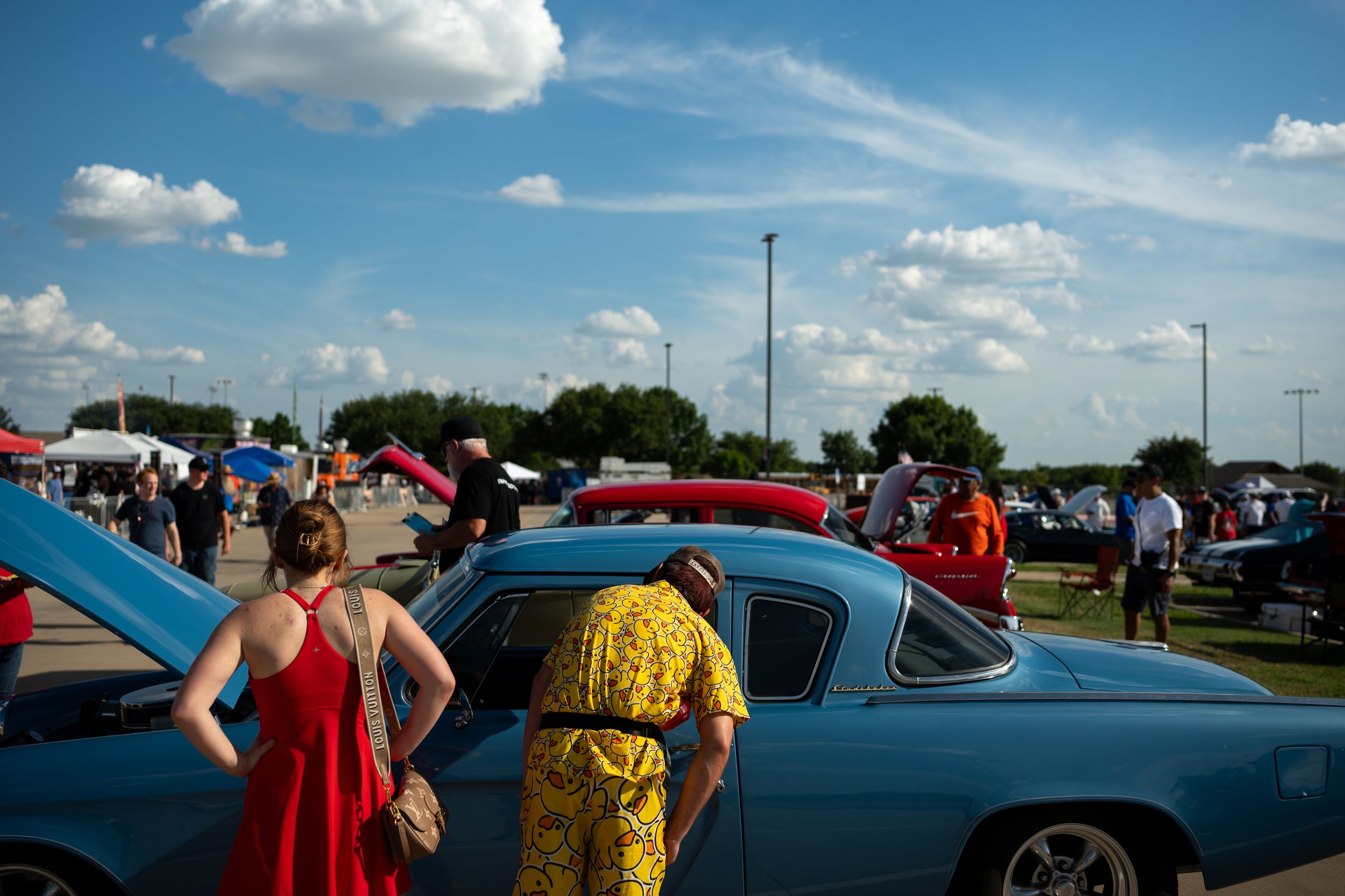 People admiring classic vintage cars at an outdoor car show on a sunny day with blue skies.