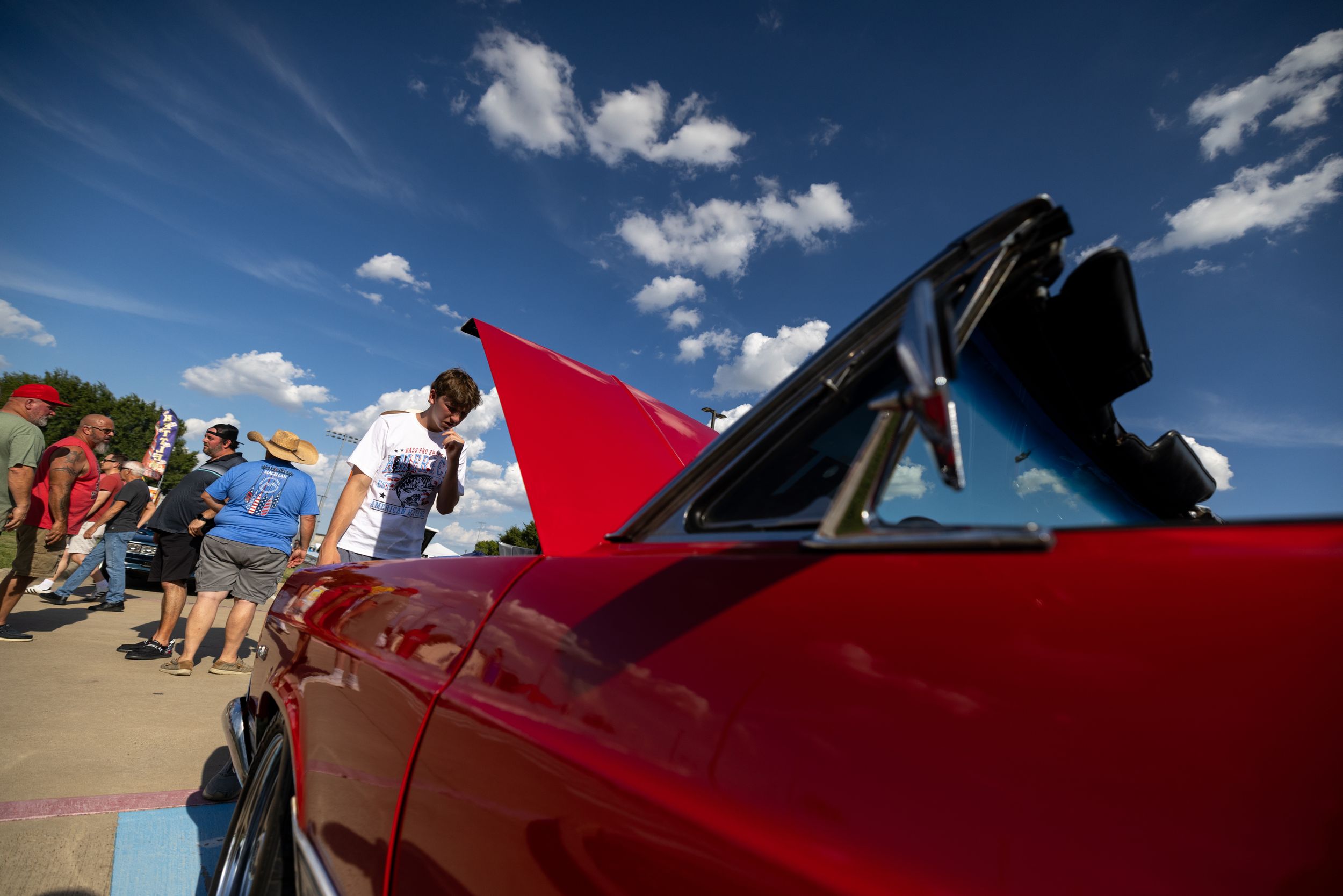 Red convertible classic car at outdoor show with man holding red flag under blue sky with clouds.