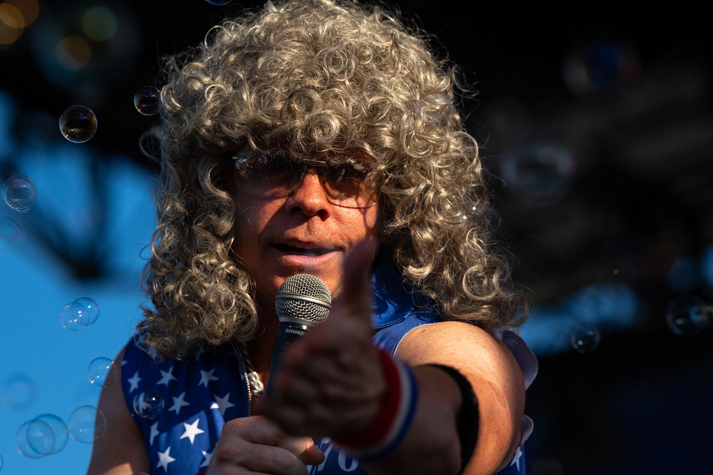 Performer wearing curly gray wig and sunglasses sings into microphone on outdoor stage with bubbles floating around.