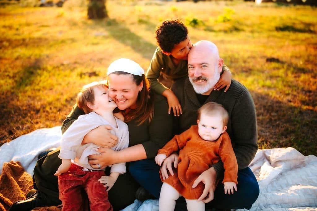 Happy family of five sits on a blanket in a sunlit autumn field, sharing kisses and warm embraces.