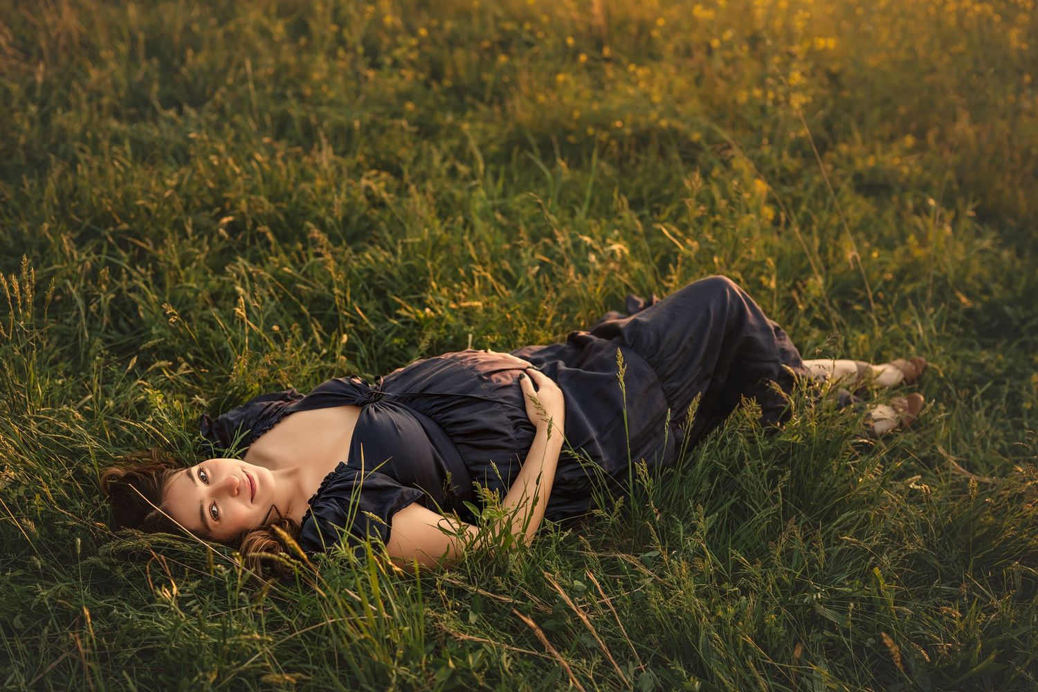 Woman in dark outfit lying relaxed in lush green meadow grass during golden hour sunset light.
