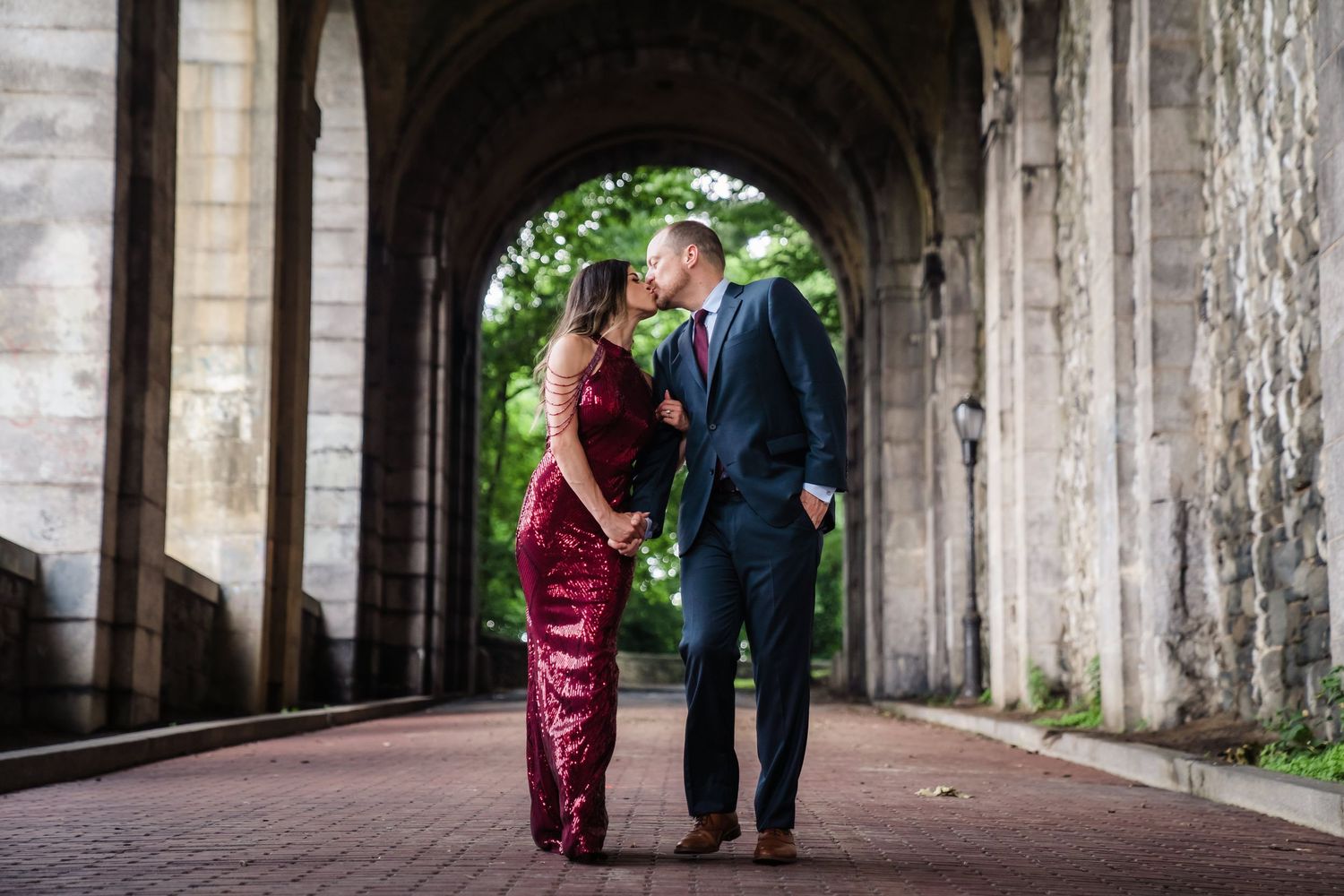 Couple in formal attire sharing a romantic moment under stone archway on brick path.
