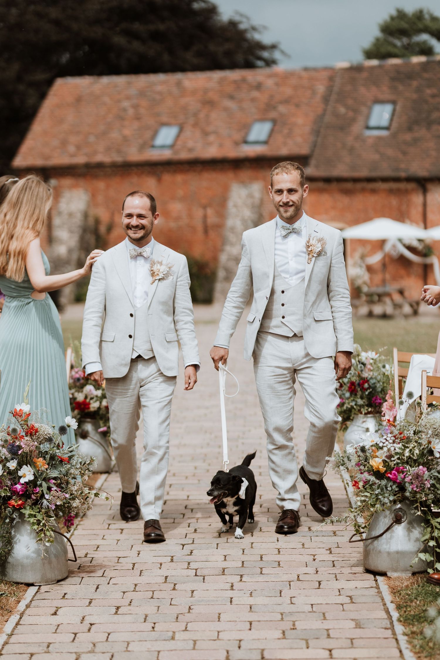 Two grooms in matching white suits walk a black dog down a floral-lined aisle at an outdoor barn wedding.