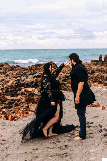 Two women in black attire lying down with dark floral wreaths adorned with red roses and ornaments on blowing rocks beach in Jupiter.