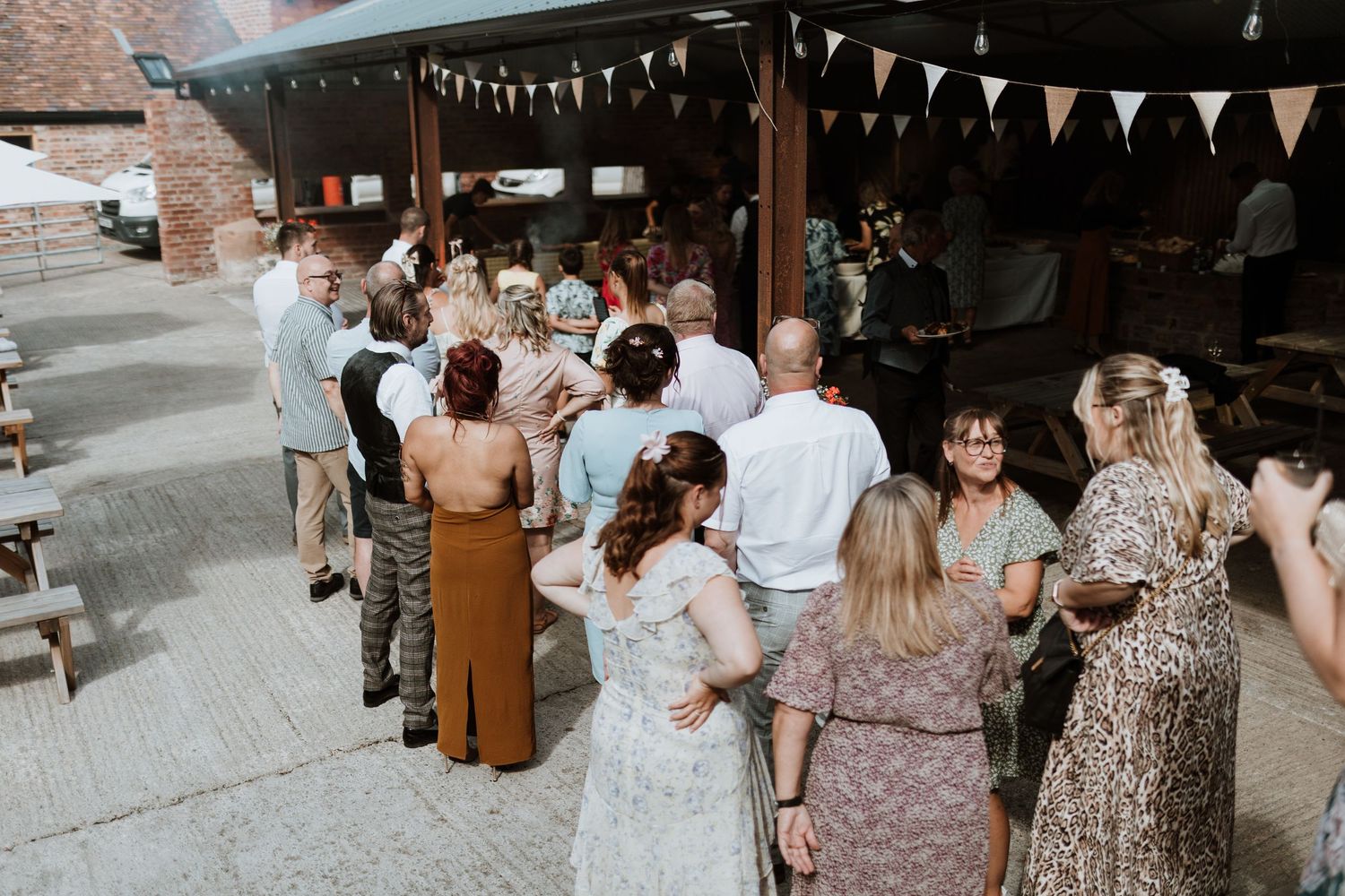Outdoor wedding reception guests mingling under bunting decorations at a rustic brick venue on a sunny day.