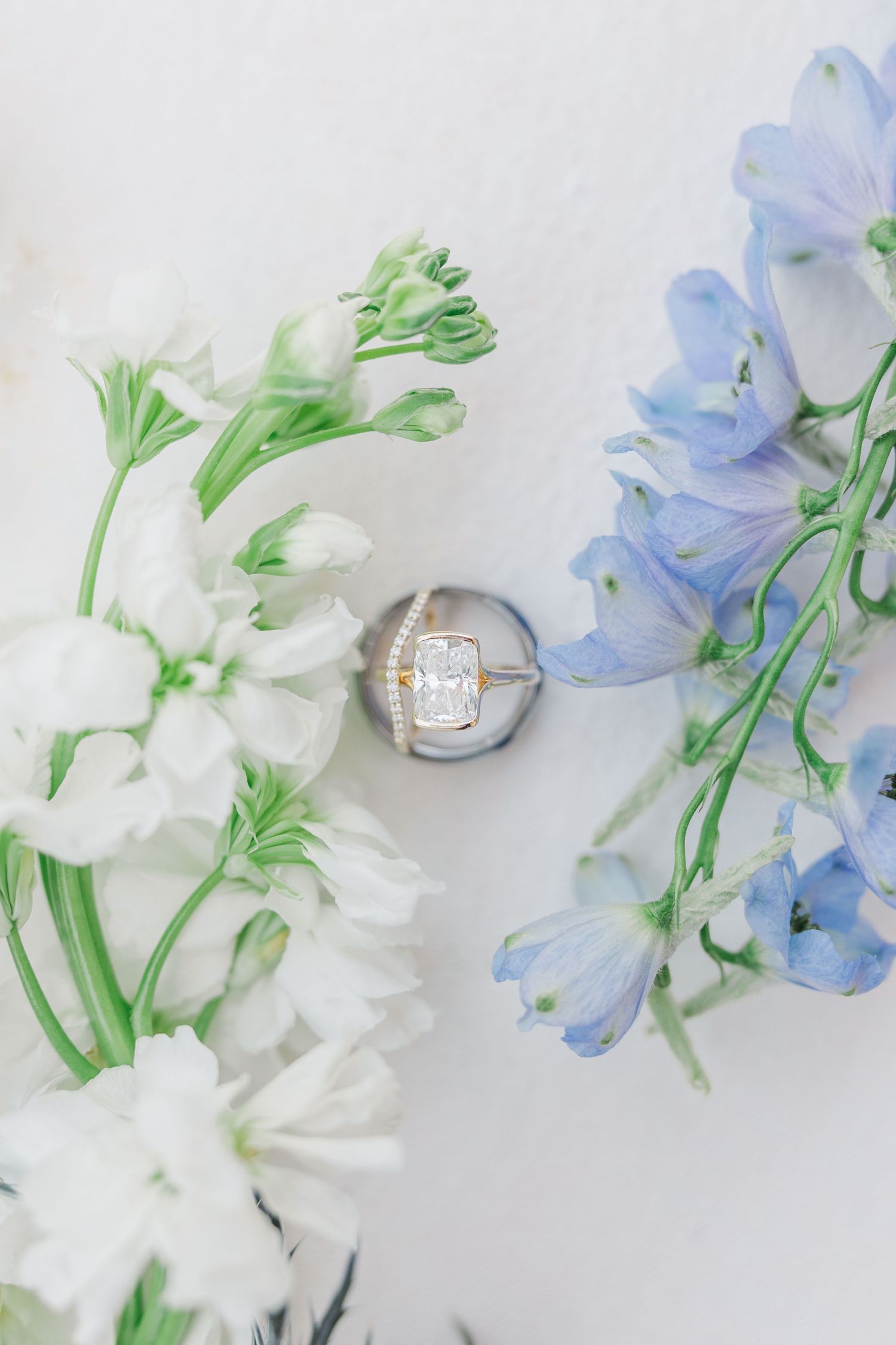 Diamond engagement ring in a silver box surrounded by white and blue delphinium flowers on a white background.