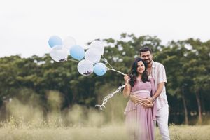 Pregnant couple holding blue and white balloons in a green field during a maternity photoshoot.