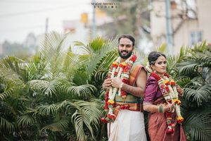 Indian couple in traditional wedding attire with floral garlands posing outdoors among tropical greenery.