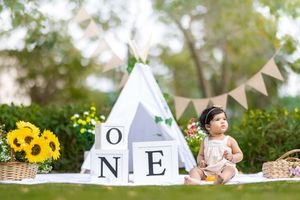 Baby girl sits outdoors near a white teepee with ONE blocks and sunflowers for a first birthday photo shoot.