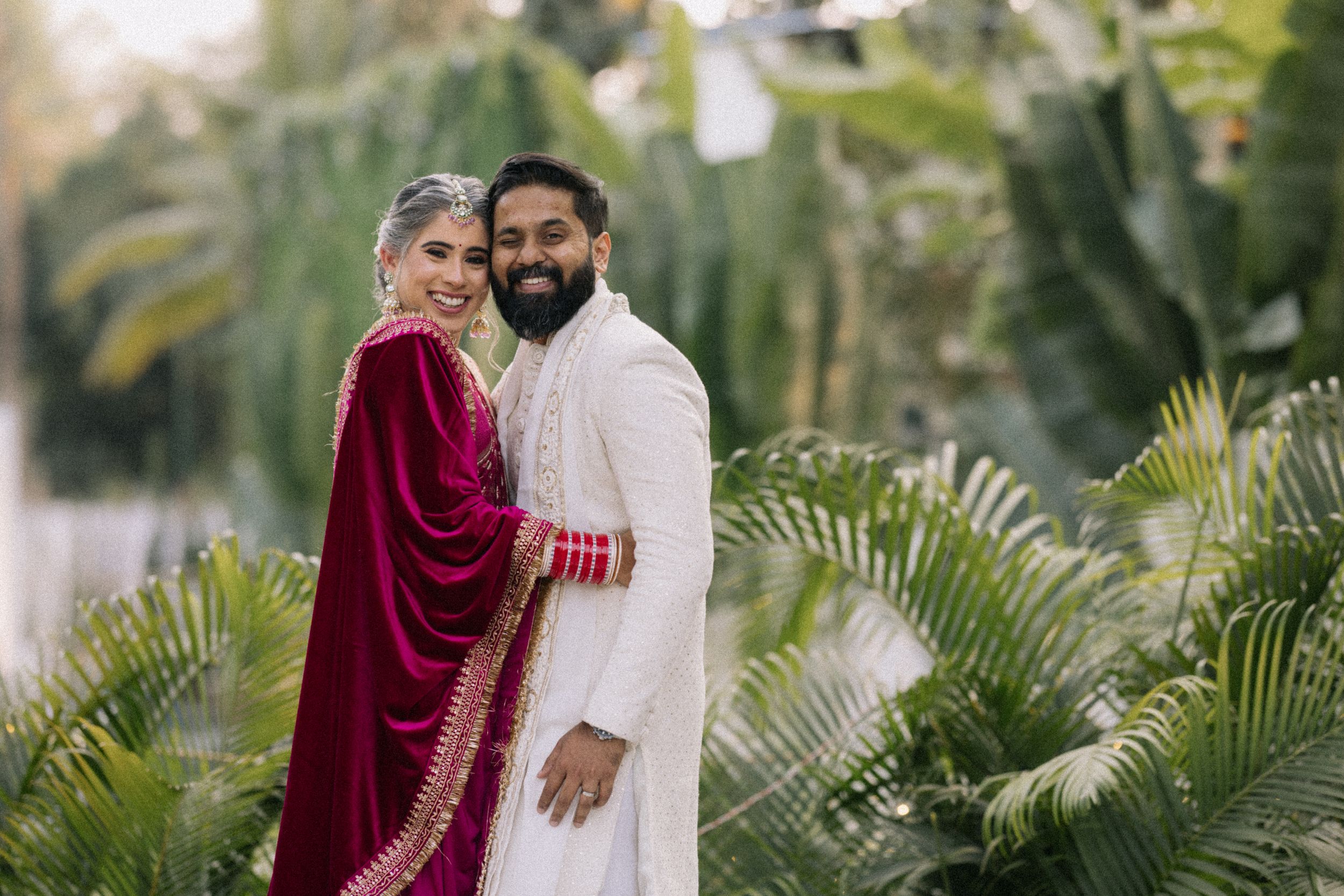 Couple in traditional South Asian wedding attire poses among lush tropical greenery, bride in red dupatta, groom in white sherwani.