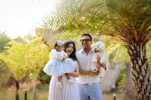 Happy family with a toddler girl and white fluffy dog posing outdoors near a palm tree in a sunny garden.