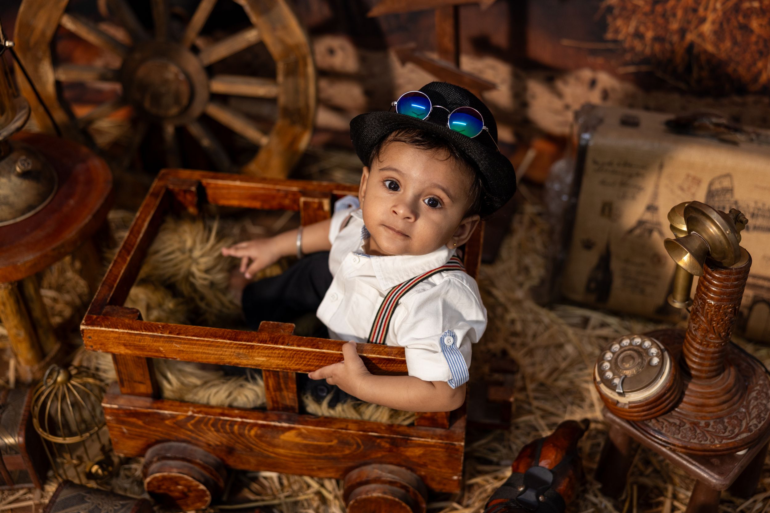 Adorable baby boy in steampunk costume with goggles and suspenders sitting in wooden toy car with vintage decor.