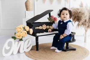 Toddler boy in navy suit sits at black toy piano with roses and a white 'One' sign for first birthday photos.