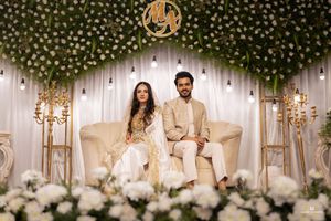 Elegant couple seated on white sofa at floral wedding backdrop with greenery arch and gold monogram decor.