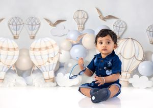Baby boy in blue outfit sits with balloons and hot air balloon backdrop in a charming cake smash photo session.