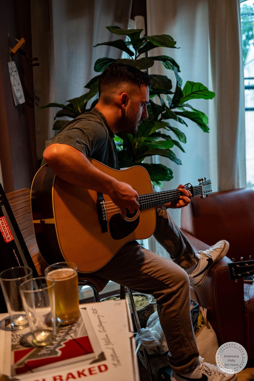 Man playing acoustic guitar while seated at an intimate indoor music venue with drinks on table.