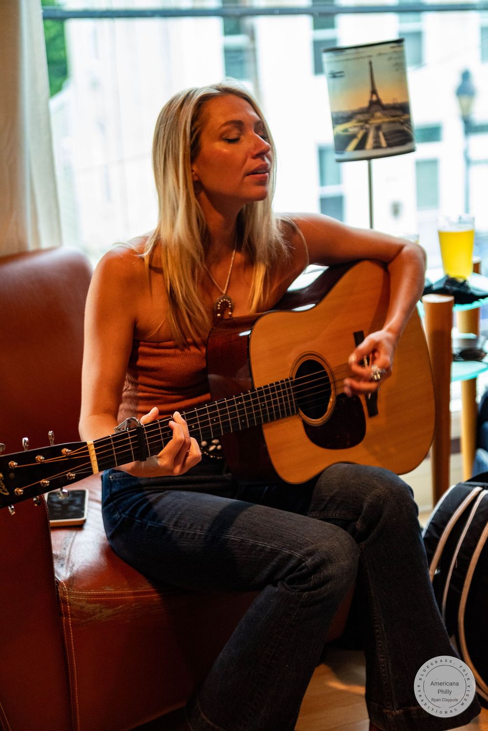 Blonde woman playing acoustic guitar while seated in a brown leather chair during an intimate indoor performance.