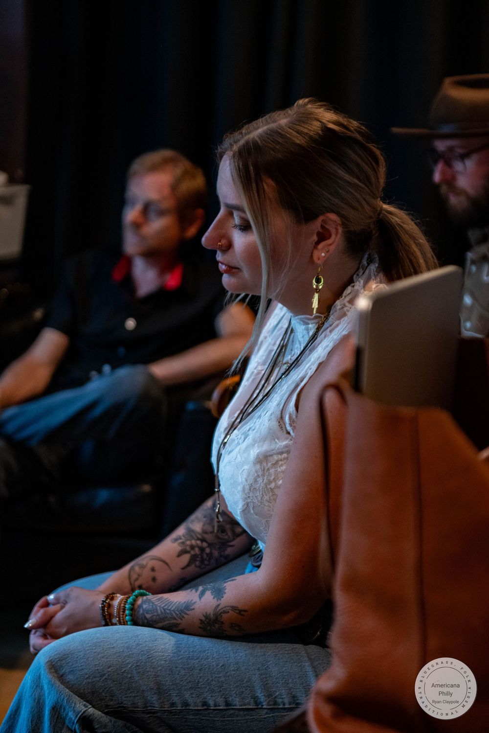 Tattooed woman with gold earrings sits attentively at an indoor event, with two people visible in the background.