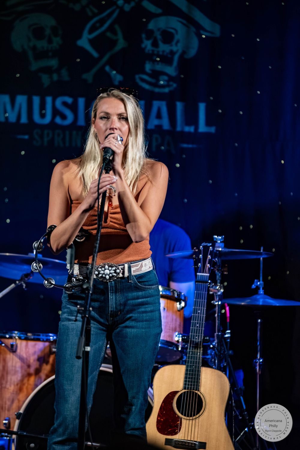 Female country singer performing on stage at Music Hall, holding microphone in orange top and jeans.