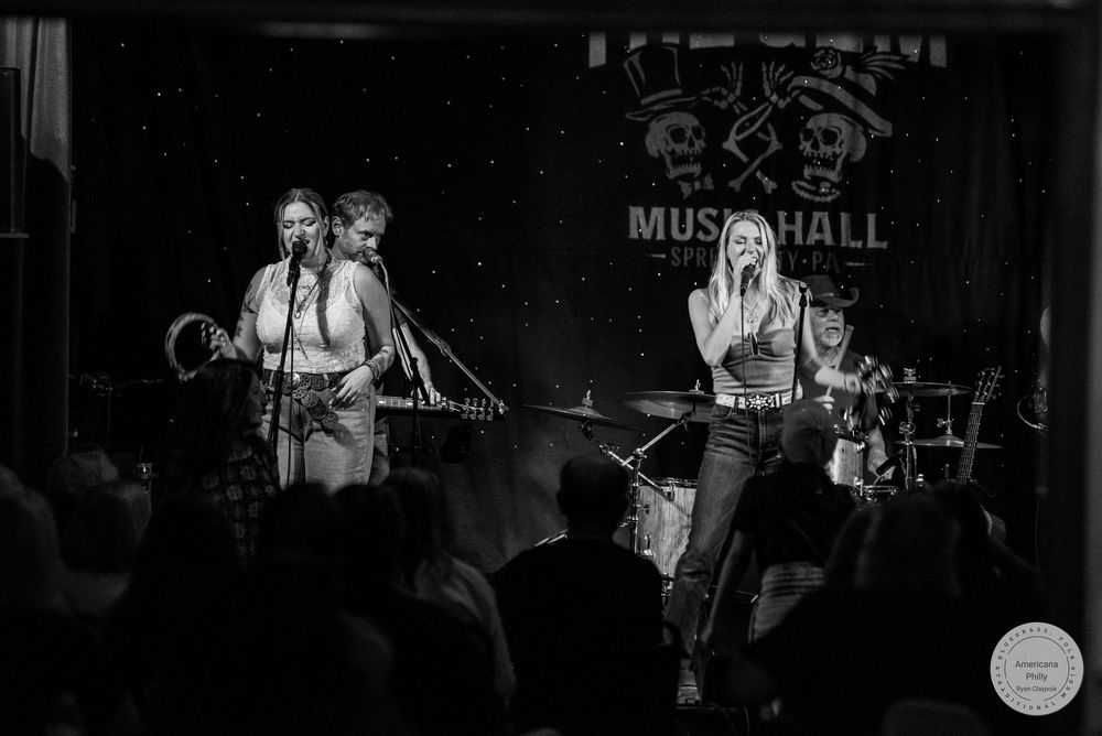 Black and white photo of a female band performing on stage at Music Hall venue with microphones and instruments.