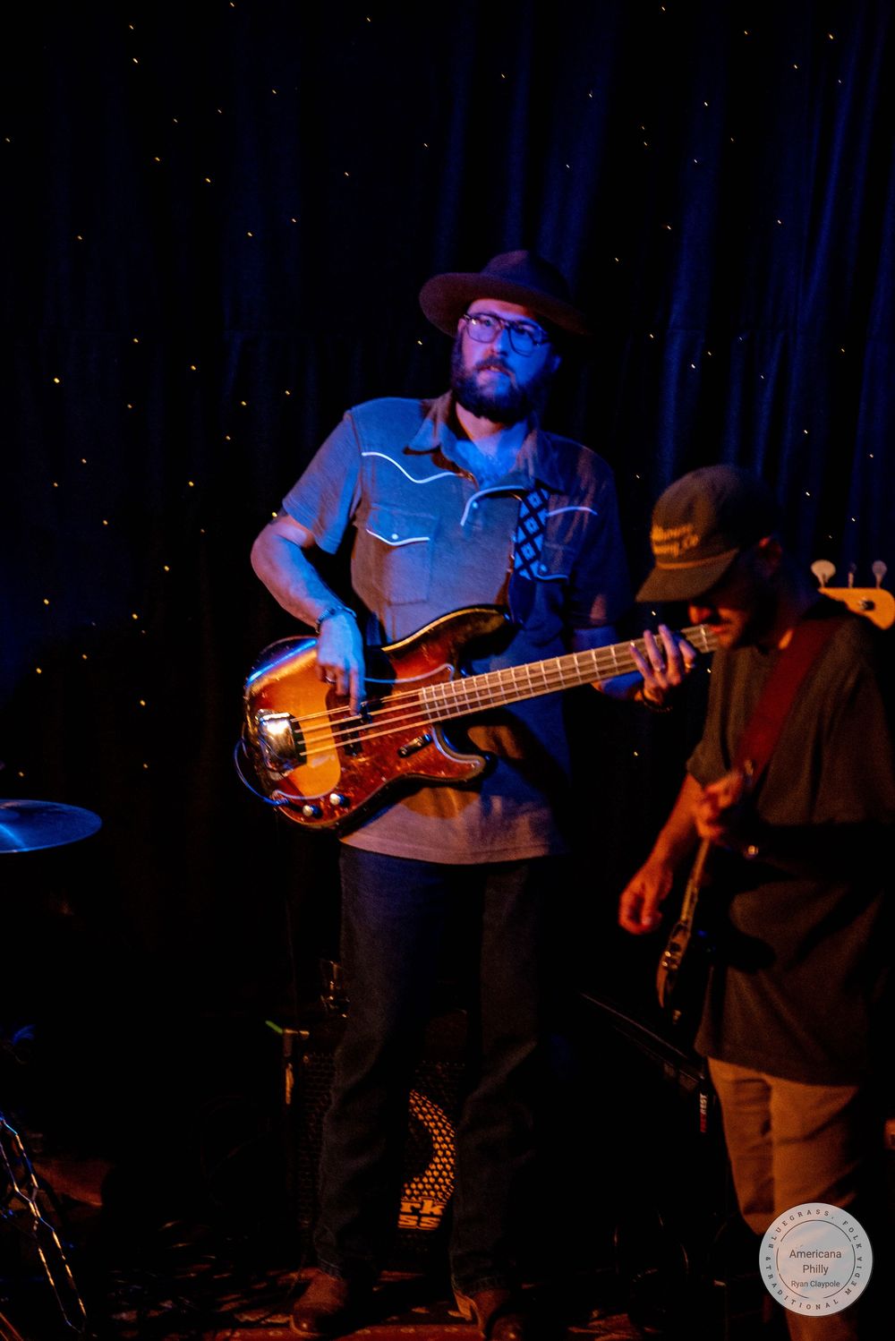 Two musicians perform on stage, one playing a bass guitar while wearing a hat under warm stage lighting.