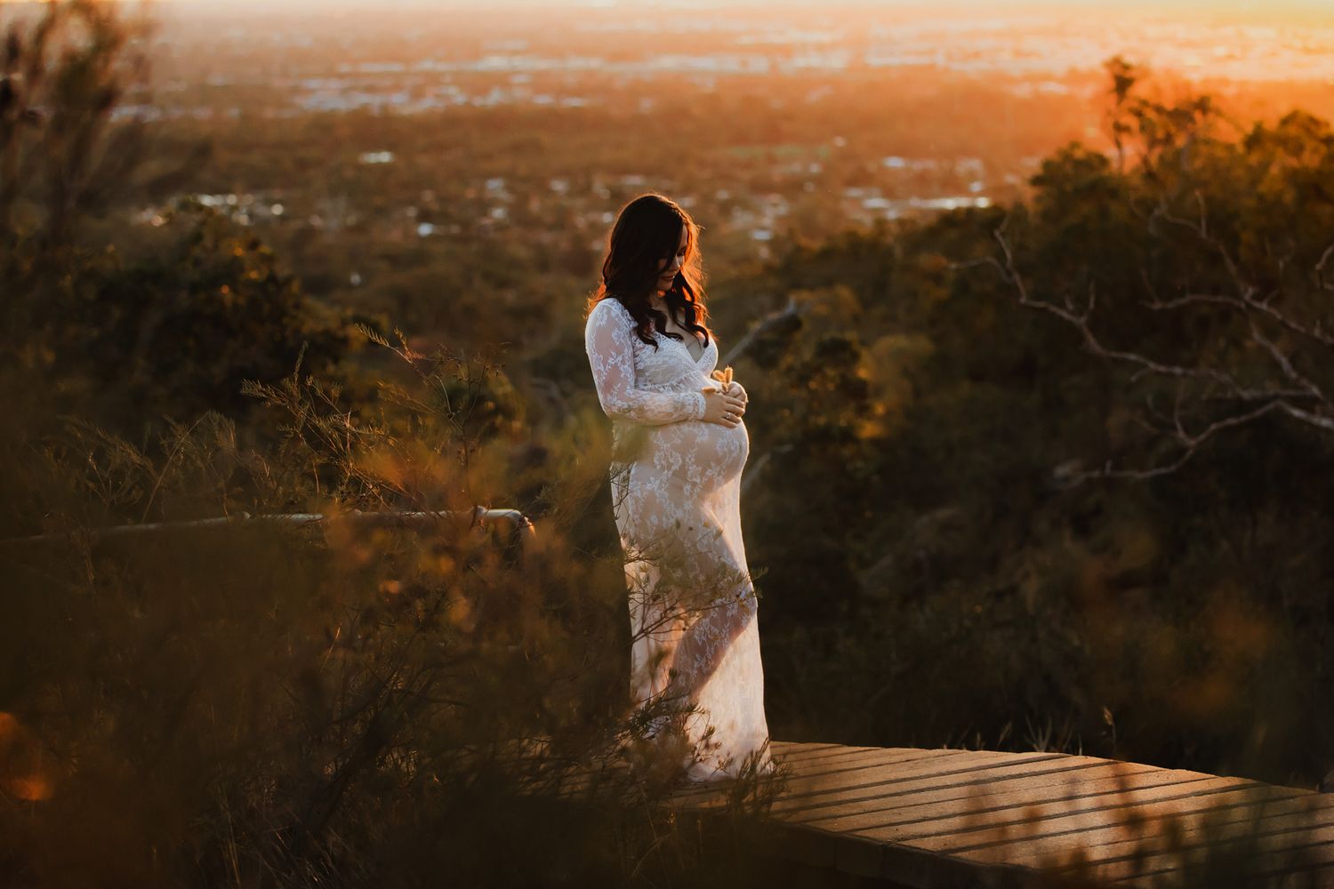 Pregnant woman in white lace dress standing on wooden deck at golden sunset with city views.