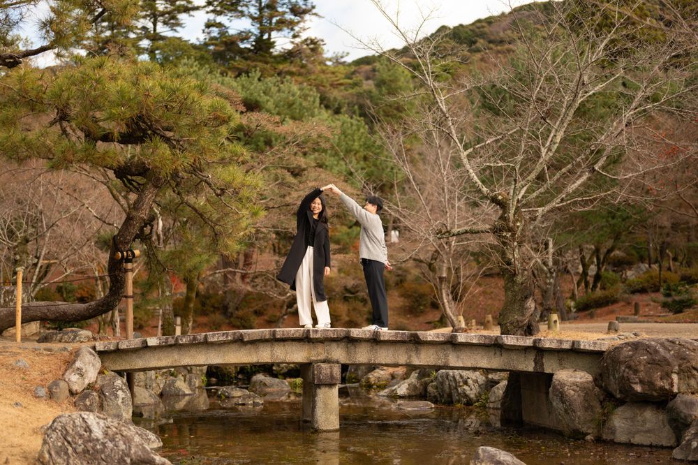 Couple holding hands and balancing on a stone bridge over a pond in a serene Japanese garden.