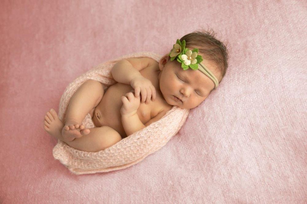 Newborn baby girl sleeping peacefully on pink blanket, wearing green flower headband, wrapped in soft pink knit wrap.