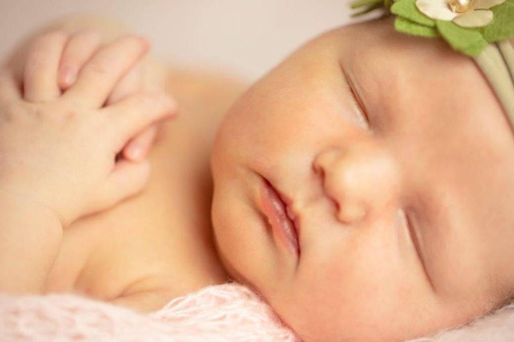 Peaceful newborn baby girl sleeping with green flower headband, soft pink background, close-up portrait photography.