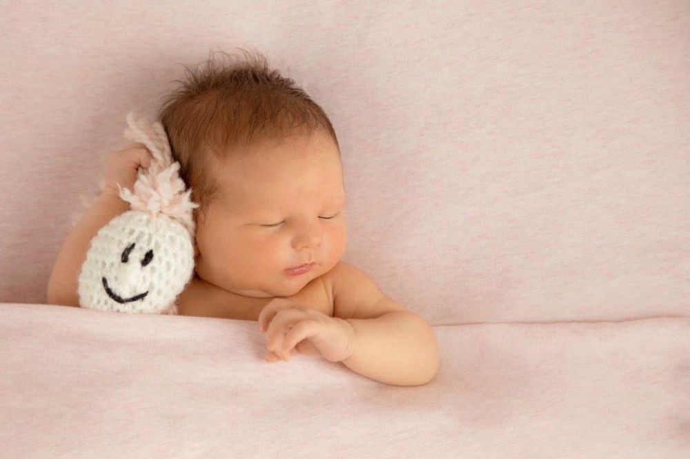 Sleeping newborn baby with pink headband resting beside a smiling knitted toy on a soft pink blanket.