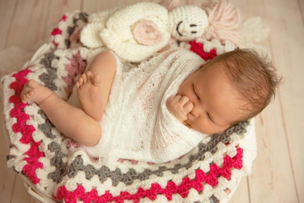Newborn baby sleeping peacefully in a crochet basket with white wrap and colorful red, gray, white blanket with stuffed toy.
