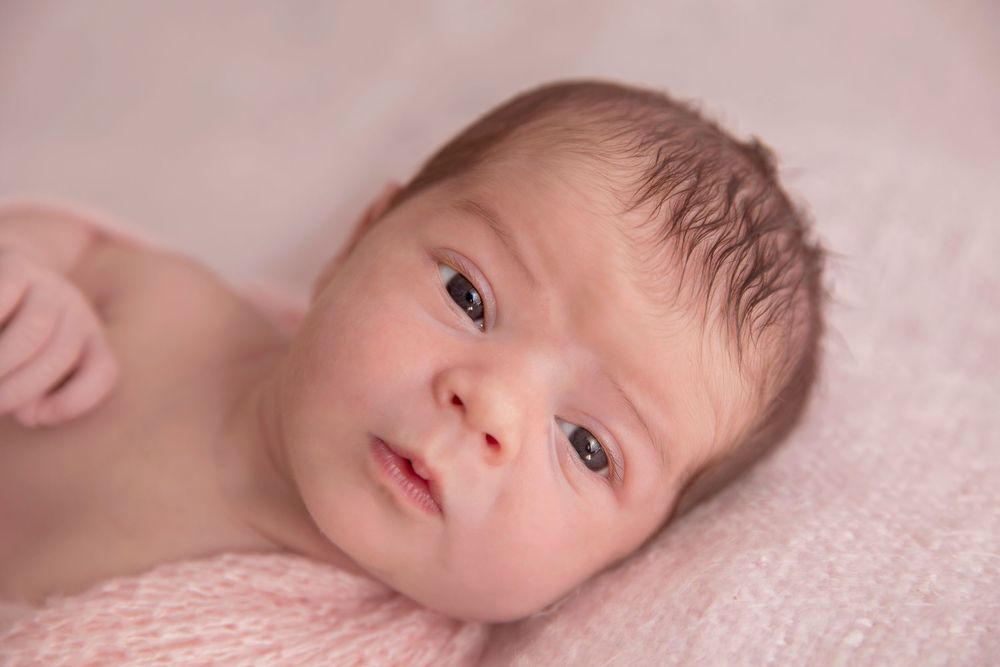 Close-up portrait of a newborn baby with dark hair lying on a soft pink blanket, eyes open and alert.