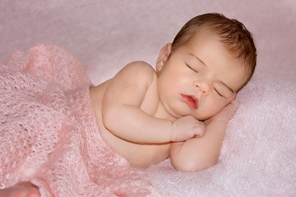 Peaceful newborn baby sleeping on a soft pink blanket, wrapped in a delicate pink knit wrap.