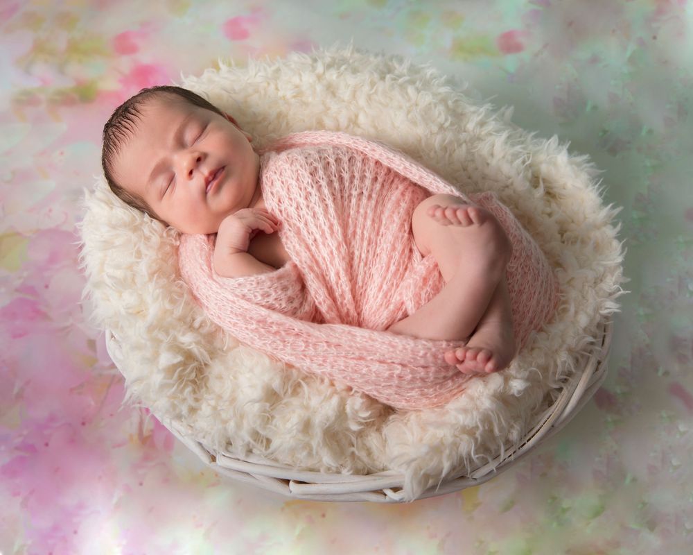 Newborn baby wrapped in pink knit swaddle sleeping peacefully in a white wicker basket with fluffy white lining.