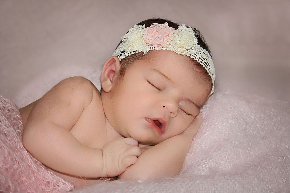 Peaceful newborn baby girl sleeping with floral lace headband on pink soft blanket.