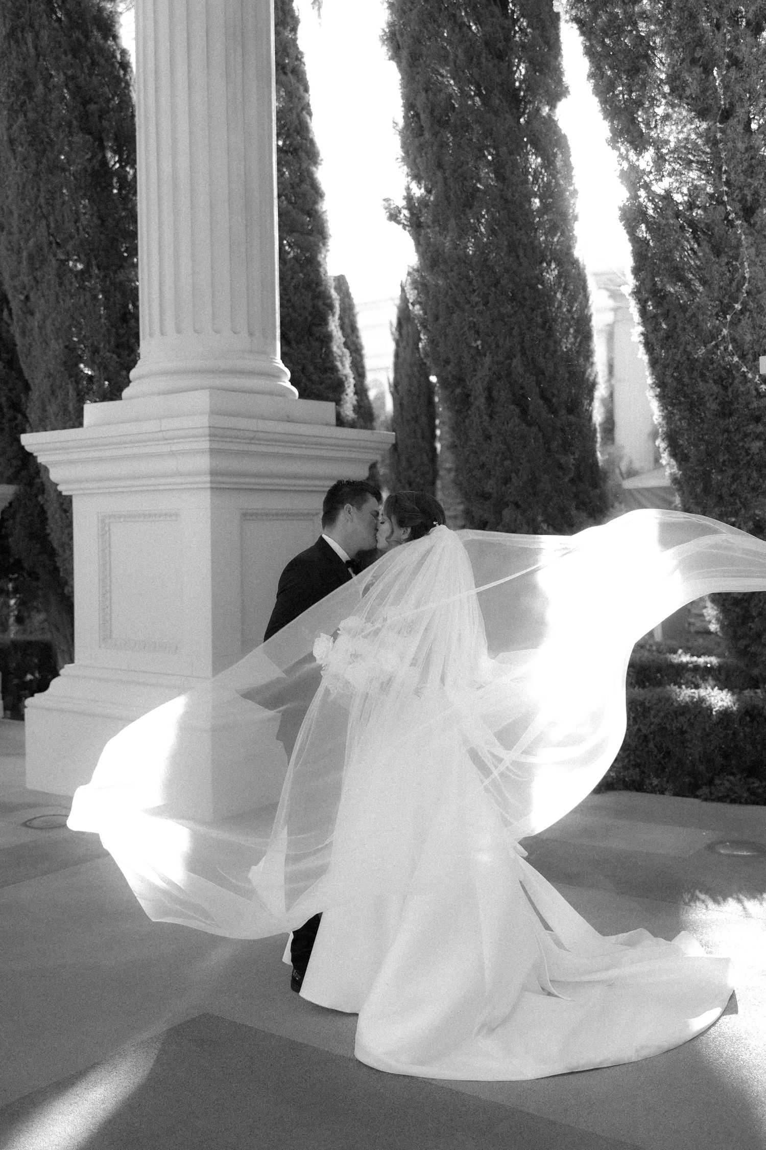 Black and white photo of a bride and groom walking outdoors, her long lace veil billowing in the wind.