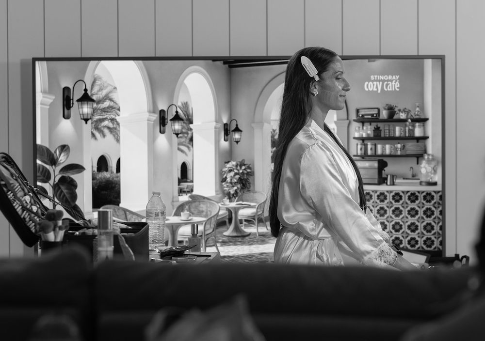 Black and white photo of a woman in a robe watching a colorful TV screen showing a cozy cafe scene.