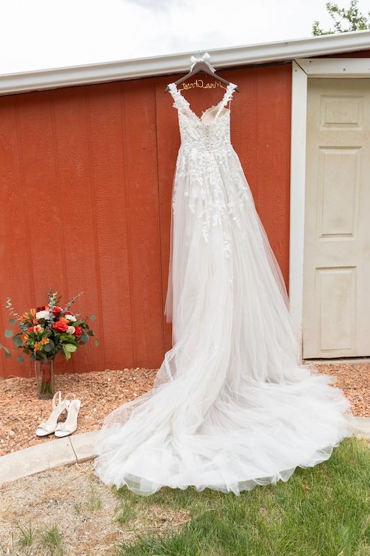 White lace wedding gown hanging outside red barn with bridal bouquet and white heels on the ground.