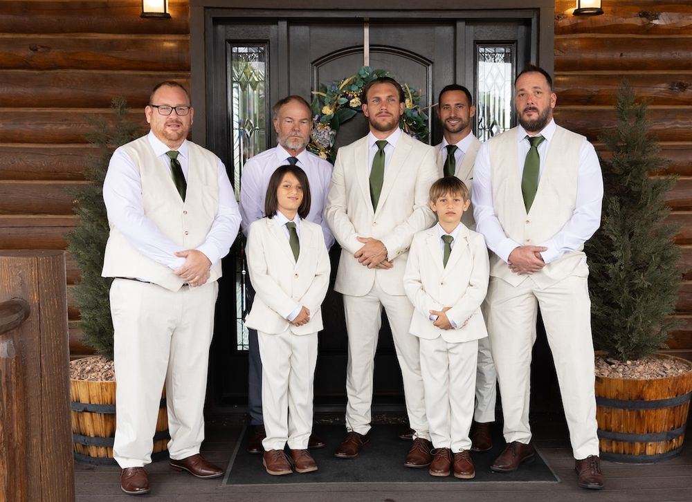 Groomsmen and ring bearers in white suits with green ties pose on rustic log cabin porch for wedding photo.