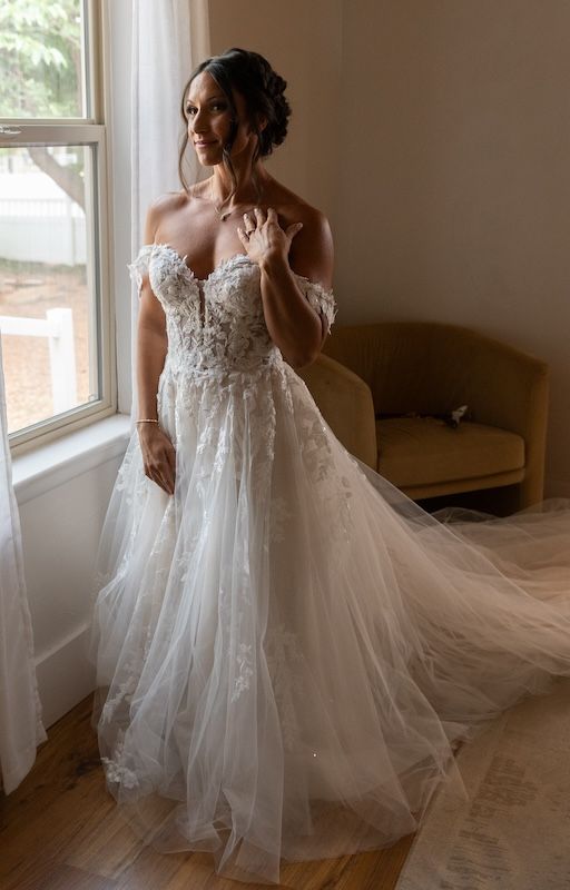 Bride in off-shoulder lace A-line wedding gown standing by window in soft natural light.